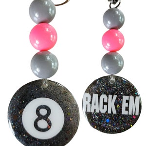 May include: A black and silver keychain with a glittery 8-ball charm and a charm that says "Rack 'Em". The keychain has three grey beads and two pink beads.