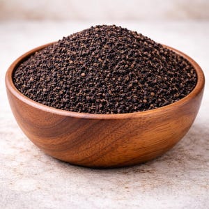 Può includere: Una ciotola di legno piena di materiale granulare marrone scuro, possibilmente foglie di tè o un prodotto secco simile. La ciotola è in legno lucido, che mostra la sua venatura naturale. Lo sfondo è una superficie strutturata neutra.