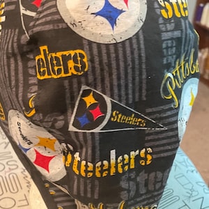 May include: A black and grey patterned cap featuring the Pittsburgh Steelers logo and text. The cap has a repeating pattern of the team's logo and the word "Steelers". The cap has a black strap.