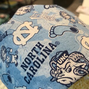 May include: Light blue fabric with a repeating pattern of white logos, text, and illustrations associated with the University of North Carolina. The words "North Carolina" and "UNC" are visible.