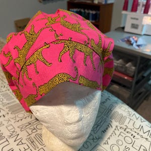 May include: A scrub cap featuring a vibrant hot pink background with a repeating pattern of yellow and black spotted leopards. The cap has a gathered, rounded crown design with stitched seams and a wide band that wraps around the head area.