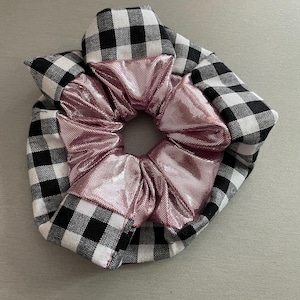 May include: A scrunchie featuring a black and white gingham pattern and a pink metallic centre. The scrunchie is made of fabric and is designed to hold hair in place. A hair accessory.