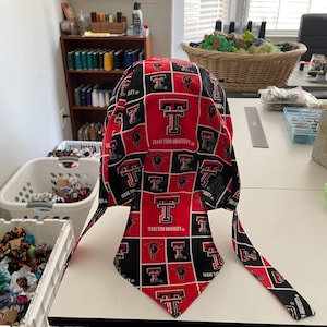 Puede incluir: Un bandana rojo y negro con el logotipo de la Universidad Tecnológica de Texas y las letras "T" y "TTU" repetidas en todo el diseño. El bandana tiene dos lazos largos.