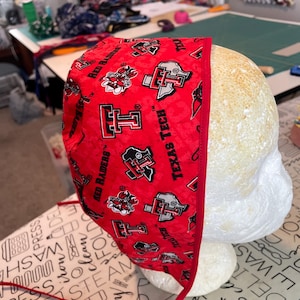 Può includere: Un cappuccio chirurgico rosso Texas Tech Red Raiders con un motivo ripetuto del logo e del testo della squadra. Il cappuccio è in tessuto e ha un bordo e dei lacci rossi.