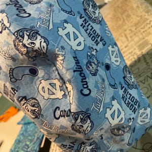 May include: Light blue fabric item with a repeating pattern of white logos, the words "UNC", "Tar Heels", and "North Carolina" are visible. The item appears to be a hat or a similar accessory.