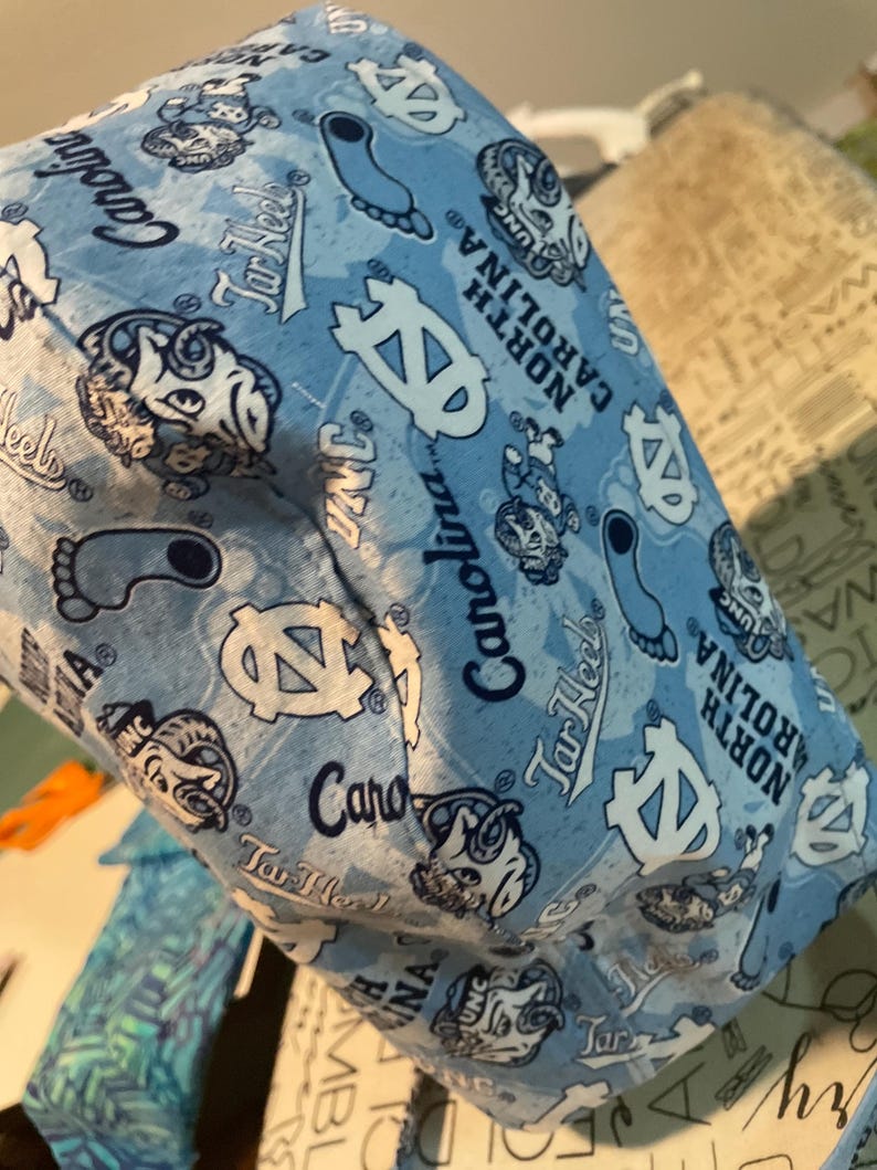 May include: Light blue fabric item with a repeating pattern of white logos, the words "UNC", "Tar Heels", and "North Carolina" are visible. The item appears to be a hat or a similar accessory.