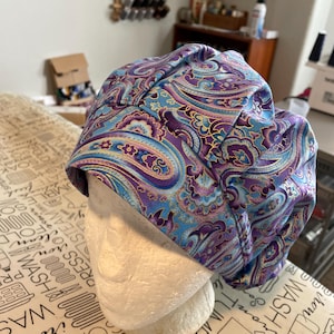 May include: A patterned surgical scrub cap in shades of blue, purple, and gold, displayed on a white mannequin head. The fabric features a paisley design. The cap is designed to cover the head and hair during medical procedures.