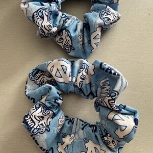 Pode incluir: Duas scrunchies azuis claras com um padr&atilde;o repetido de logotipos e texto brancos. As scrunchies s&atilde;o feitas de tecido e s&atilde;o franzidas para criar um efeito de babado. Os logotipos e o texto s&atilde;o em estilo universit&aacute;rio.