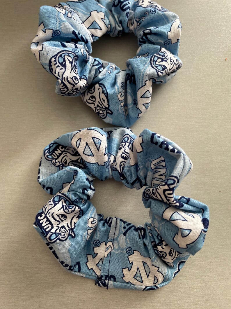 Pode incluir: Duas scrunchies azuis claras com um padr&atilde;o repetido de logotipos e texto brancos. As scrunchies s&atilde;o feitas de tecido e s&atilde;o franzidas para criar um efeito de babado. Os logotipos e o texto s&atilde;o em estilo universit&aacute;rio.