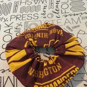 Op de afbeelding: Een bordeauxrode en gouden scrunchie met de tekst "WASHINGTON COMMANDERS" erop gedrukt. De scrunchie is gemaakt van stof en heeft een cirkelvorm. De achtergrond is een witte stof met wasgerelateerde tekst en afbeeldingen.