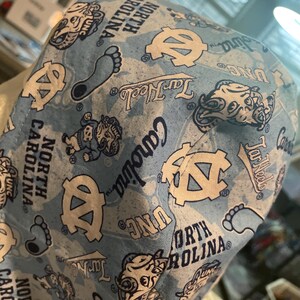 May include: Light blue fabric with a pattern of UNC logos, Tar Heels text, and the words "North Carolina." The design includes the UNC logo, a foot, and a cartoon mascot. The fabric is likely used for a hat or similar item.