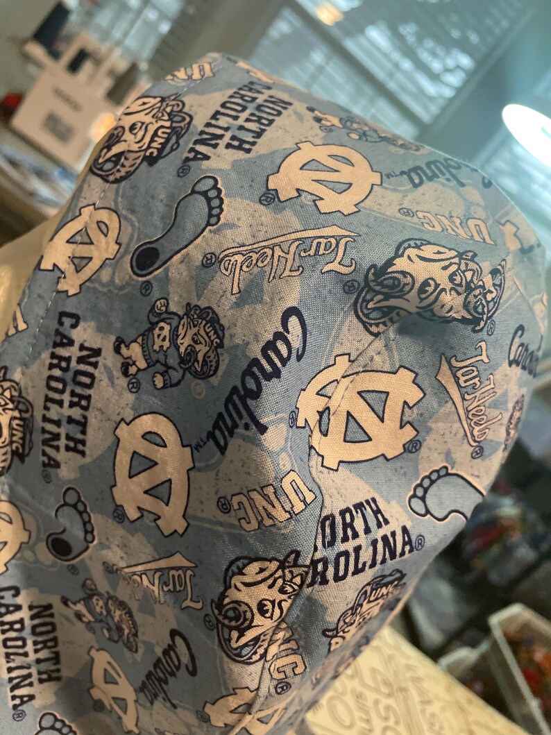 May include: Light blue fabric with a pattern of UNC logos, Tar Heels text, and the words "North Carolina." The design includes the UNC logo, a foot, and a cartoon mascot. The fabric is likely used for a hat or similar item.