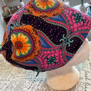 May include: A patterned surgical scrub cap. The cap has a dark background with bright floral and paisley designs in orange, pink, blue, and green. The cap is displayed on a white mannequin head.