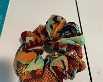May include: A patterned scrunchie in shades of orange, teal, and burgundy. The fabric has a geometric design. The scrunchie is circular and likely made from a soft material, ideal for hair styling.