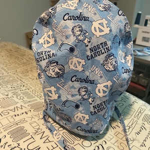 May include: A light blue surgical scrub cap featuring a repeating pattern of white logos, text, and cartoon figures. The text includes "UNC", "Carolina", and "Tar Heels". The cap has ties for securing and is made of fabric.