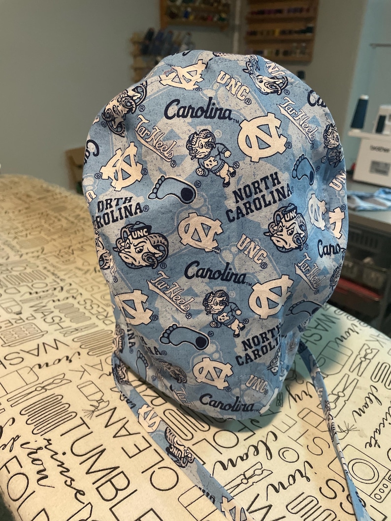 May include: A light blue surgical scrub cap featuring a repeating pattern of white logos, text, and cartoon figures. The text includes "UNC", "Carolina", and "Tar Heels". The cap has ties for securing and is made of fabric.