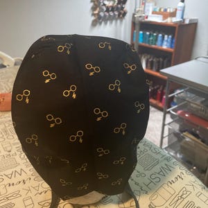 May include: A black scrub cap featuring a repeating gold pattern of round eyeglasses and lightning bolts. The fabric is dark with metallic gold icons distributed evenly across the surface, and the cap includes black fabric ties at the base.