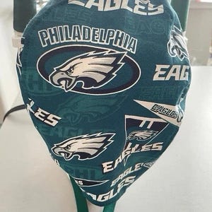 Könnte beinhalten: Eine blaugrüne und weiße Philadelphia Eagles OP-Haube. Die Haube zeigt das Logo des Teams, einen Adlerkopf, und die Worte "EAGLES" und "PHILADELPHIA" in Weiß. Die Haube ist aus Stoff und hat ein grünes Band.