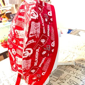 May include: A red and white patterned cap featuring the text "OKLAHOMA SOONERS" and various related graphics. The cap has a red ribbon tie and is likely made of fabric. The design includes the words "BOOMER SOONER".