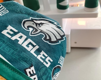 May include: A teal and white Eagles print fabric is the focal point of this image. The fabric is made into a cap with the word "EAGLES" in white block letters and a white and black eagle logo. The cap has a green trim.