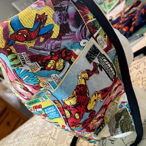 May include: A Marvel comic book print fabric tote bag featuring Spider-Man, Iron Man, and other characters. The bag has a black trim and straps. The comic book panels are in bright colors, including red, yellow, blue, and purple.