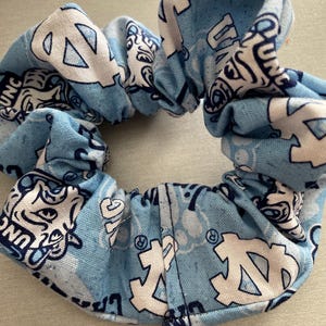 Pode incluir: Uma scrunchie azul claro com um padr&atilde;o repetido de logotipos e texto brancos. A scrunchie &eacute; feita de tecido e &eacute; reunida em torno de uma faixa el&aacute;stica. O design apresenta as letras "UNC" e um logotipo estilizado.