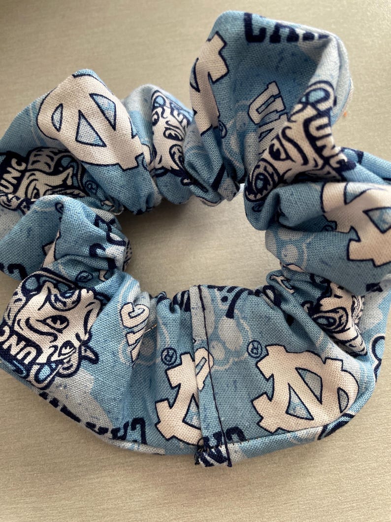 Pode incluir: Uma scrunchie azul claro com um padr&atilde;o repetido de logotipos e texto brancos. A scrunchie &eacute; feita de tecido e &eacute; reunida em torno de uma faixa el&aacute;stica. O design apresenta as letras "UNC" e um logotipo estilizado.
