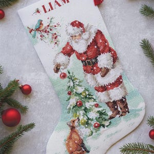 Può includere: Una calza di Natale bianca con un disegno a punto croce raffigurante Babbo Natale che decora un albero di Natale. La calza ha il nome "LIAM" in lettere rosse in alto. Una volpe è alla base dell'albero. Ornamenti rossi e verde circondano la calza.