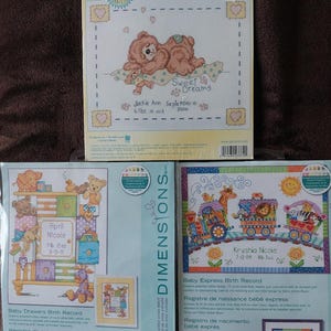 May include: Three cross-stitch kits for baby birth records. The top kit features a bear design with the text "Sweet Dreams." The other two kits show a baby drawers design and a train design. Each kit includes the dimensions in cm.