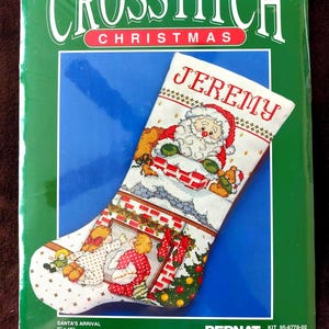 May include: A Christmas cross-stitch kit with a stocking design. The white stocking features a Santa Claus motif, the name "JEREMY" in red, and other festive figures. The kit includes fabric, thread, and instructions. The package is green with the words "CROSSSTITCH CHRISTMAS". The stocking measures 23 cm x 41 cm.