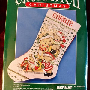 May include: A Bernat Crosstitch Christmas kit featuring a 9 inch by 16 inch stocking design with bears and reindeer. The packaging displays the name Carrie, the text Merry Christmas, and lists contents including 14 count aida fabric and floss.