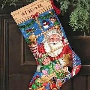May include: Christmas stocking with a festive design featuring Santa Claus, children, and toys. The stocking is primarily blue, red, and green, with the name "ABIGAIL" at the top. A red tassel hangs from the top.
