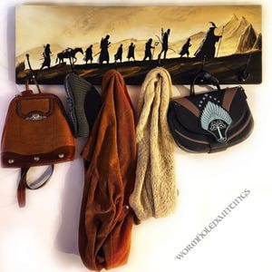 May include: A wooden wall-mounted coat rack with a painted image of the Lord of the Rings characters walking across a landscape. There are four hooks on the rack, with a brown leather backpack, a gray knitted hat, a brown corduroy jacket, a beige knitted scarf, and a black leather bag with a silver tree design hanging from the hooks. The text "wormholepaintings" is visible in the bottom right corner of the image.