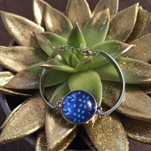 Puede incluir: Un brazalete de plata con un cabujón de vidrio azul y blanco con estampado. El brazalete está sobre un fondo de purpurina dorada.