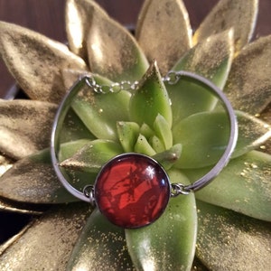 Op de afbeelding: Een zilveren armband met een rode glazen cabochon. De armband ligt op een groene succulent met gouden glitter.