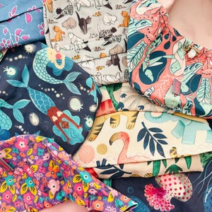 May include: A collection of colorful fabric items with various patterns. Designs include mermaids, cats, flowers, flamingos, and abstract shapes. The color palette is diverse, featuring blues, pinks, purples, and greens. The items appear to be handmade.