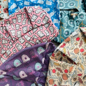 May include: A collection of fabric scrub caps featuring various patterns including white ghosts on blue, white ghosts on pink, purple jellyfish-like shapes, teal floral designs, and a cream background with autumn pumpkins, leaves, and pretzels.
