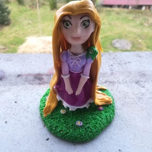 Puede incluir: Figura artesanal de una princesa con largo cabello rubio, vestida con un vestido morado con cuello blanco. La figura está sentada sobre una base verde con pequeñas flores. Un camaleón verde está en su hombro.