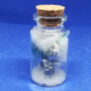 Peut inclure: Petite bouteille en verre avec bouchon en liège, contenant des figurines miniatures et de la ouate blanche. La bouteille contient un chien gris et un ours polaire blanc, sur fond bleu.