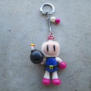 May include: A handmade Bomberman character keyring. The figure has a white helmet, blue suit, pink gloves and shoes, and holds a black bomb. The keyring has a silver ring and decorative pink and white beads.