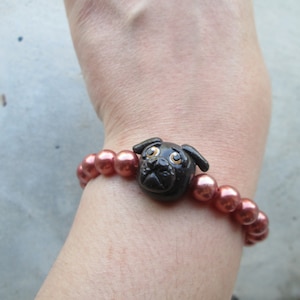May include: A bracelet with reddish-brown beads and a black pug dog charm. The charm is a detailed pug head. The bracelet is worn on a wrist. The beads are strung together to form a piece of jewellery.