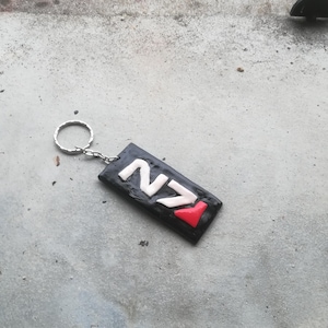 May include: A black and white keychain with the letters "N7" in white and a red arrow pointing to the right. The keychain is attached to a silver metal ring.