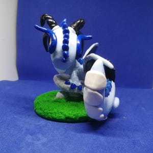 Puede incluir: Figura artesanal de una criatura azul y blanca con cuernos y cola. La figura está sobre una pequeña base circular verde, con un fondo azul.