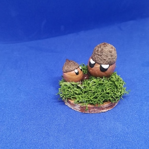 May include: Two whimsical acorn figurines with painted faces and brown caps, nestled on a bed of green moss. The base is a circular piece of wood. The background is a solid blue.