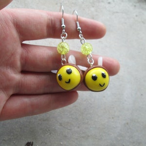 May include: A pair of dangle earrings featuring yellow and brown bee-shaped charms with white wings, black eyes, and a smiling mouth. Each earring includes a translucent lime green crackle bead attached to a silver-toned metal hook finding.