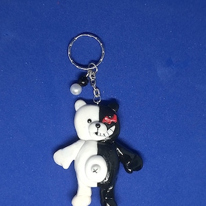 May include: A handmade keychain featuring a split-colored bear character. One half is white, the other black, with a red eye and a stitched mouth. The keychain includes a silver ring and decorative beads. The bear is approximately 2 inches tall.
