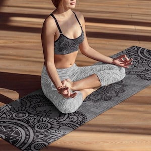 May include: A woman sits in a yoga pose on a black and white mandala patterned yoga mat. The mat has a grey background with black mandala designs.