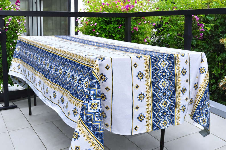 May include: White tablecloth with a blue and gold geometric pattern. The tablecloth is draped over a black metal table on a tiled patio.