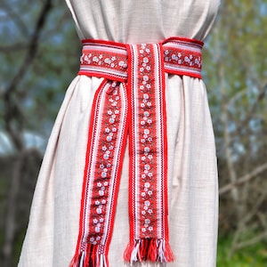 Könnte beinhalten: Ein weißes Leinenkleid mit einem roten und weißen bestickten Gürtel. Der Gürtel hat ein Blumenmuster und Quasten.