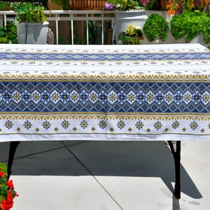 May include: White tablecloth with a blue and gold geometric pattern. The tablecloth is laid on a table with a black metal frame. The table is outdoors on a patio.